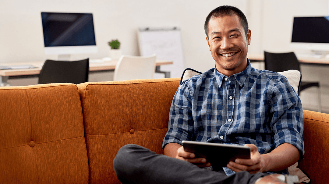 A smiling man in a plaid shirt sitting on an orange sofa in an office, holding a tablet.