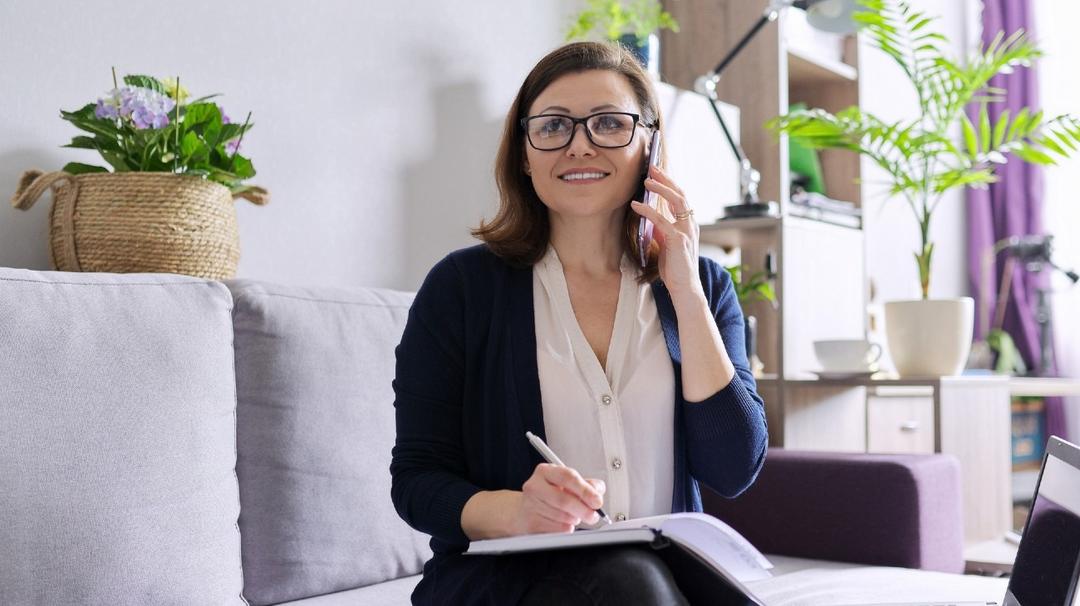 A woman in glasses, sitting on a couch, talks on a smartphone and writes in a notebook, with a laptop and indoor plants in the background.