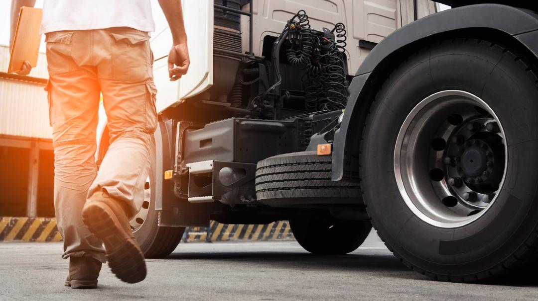 A truck driver walking towards the cab of a semi-truck in a sunny freight depot.