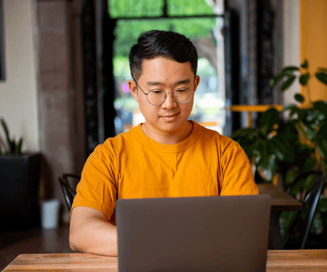 A young asian man in a yellow t-shirt working on a laptop at a cafe table, with blurred greenery in the background.